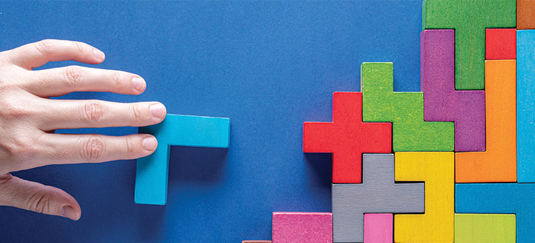 hand reaches out to place blue wooden Tetris puzzle piece into colorful stack of pieces