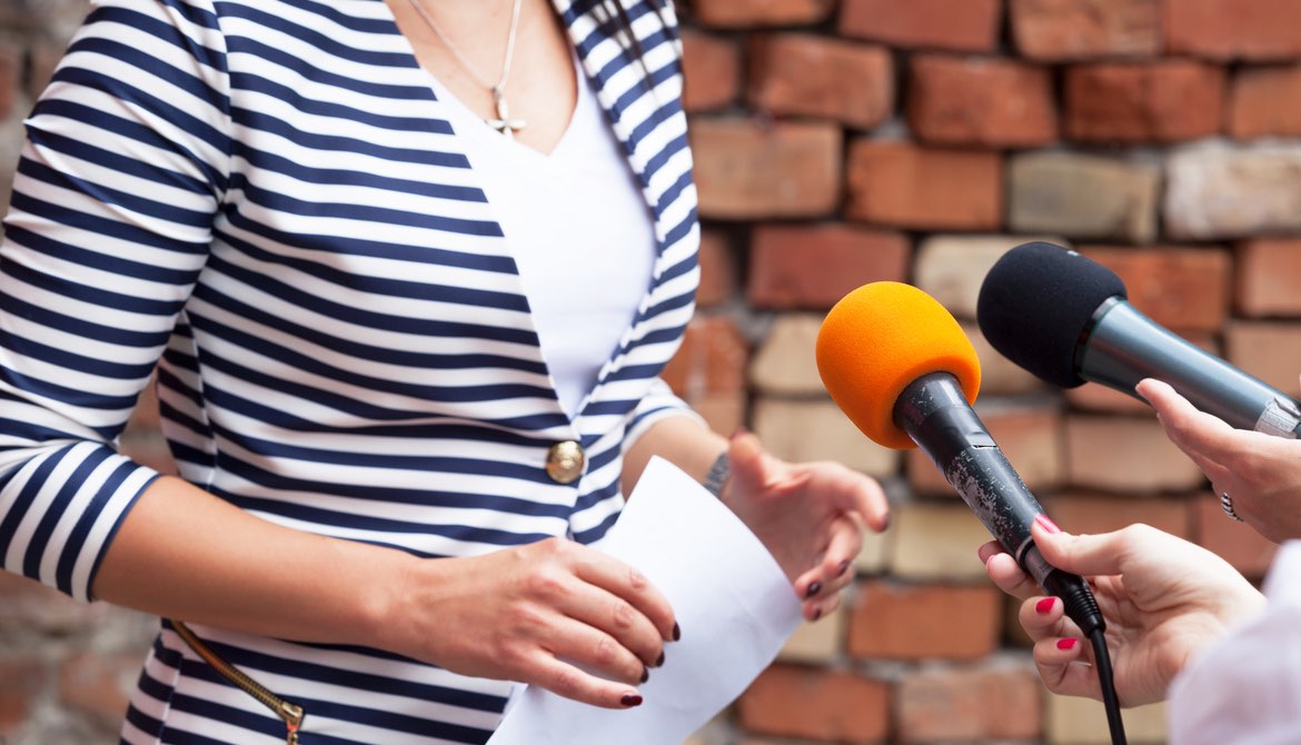 businesswoman in striped jacket speaking to interviewers with microphones