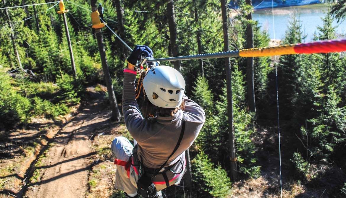 woman riding down zip line over pine trees