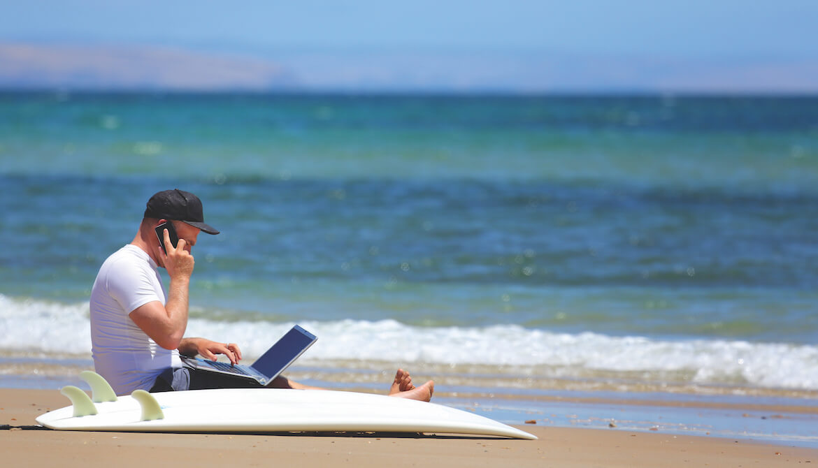 Many works or learns on a laptop at the beach next to a surf board