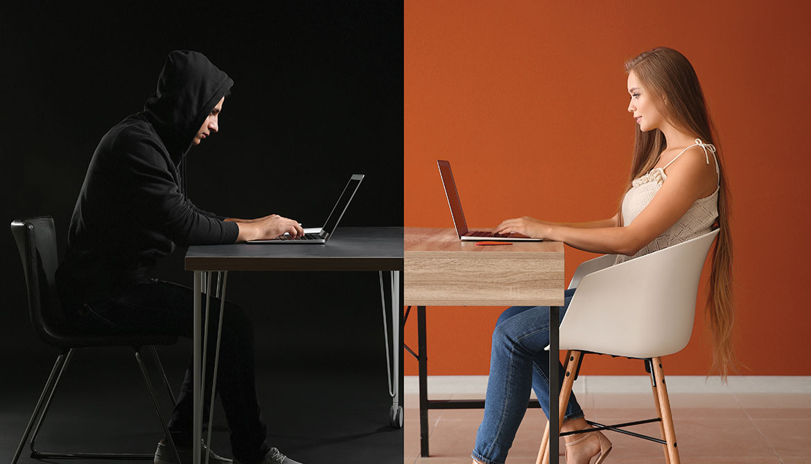 young woman working on laptop across table from a hacker wearing a dark hoodie
