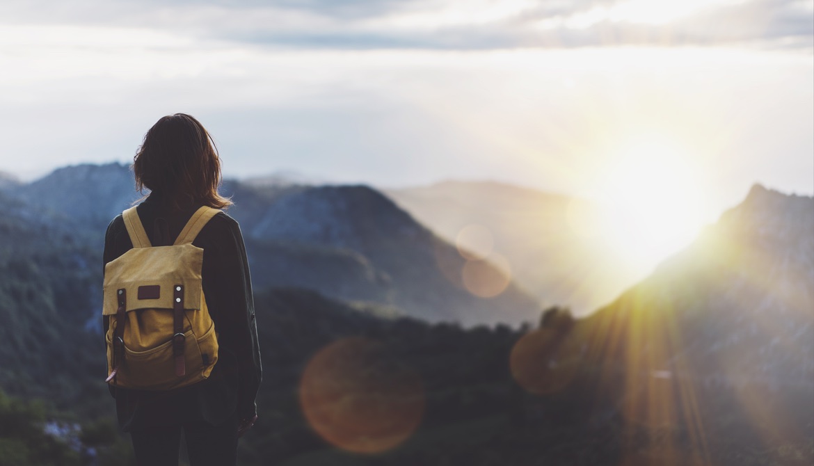 woman with backpack gazing toward sunset over mountain peaks