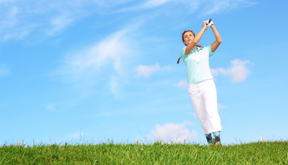 female golfer following through swing in the rough in front of blue sky