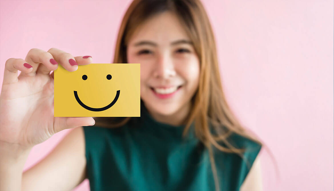 smiling young Asian woman in green shirt holds up yellow notecard with a smiley face drawn on it