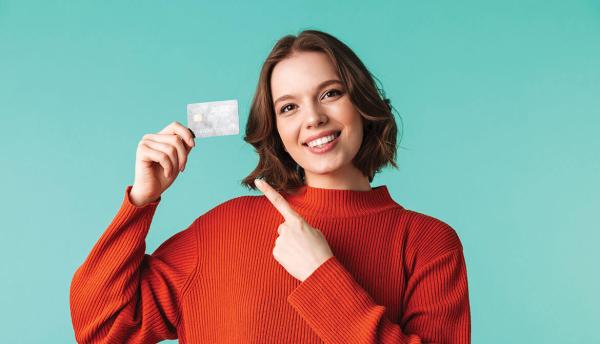Smiling young woman in orange sweater holding up and pointing to a credit card