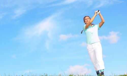 female golfer following through swing in the rough in front of blue sky