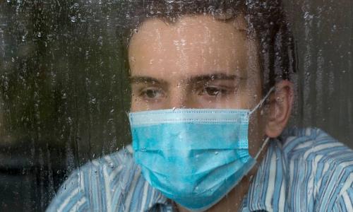 sad man wearing surgical mask looking out window in rain