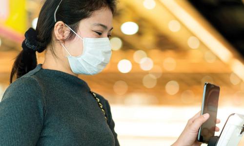 young Asian woman wearing mask holds smartphone up to payment kiosk at store