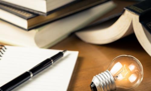 Small light bulb glowing on the desk with notebook and many books on background