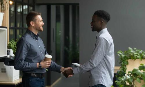 White and black male coworkers shake hands and smile at each other holding cups of coffee in an office