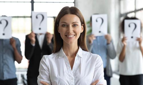 confident smiling businesswoman stands in front of row of job candidates who are holding question marks in front of their faces 