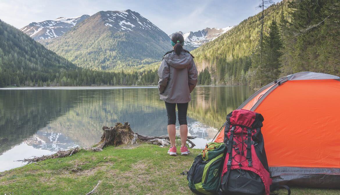 Woman at campsite looks over a lake to a distant mountain peak