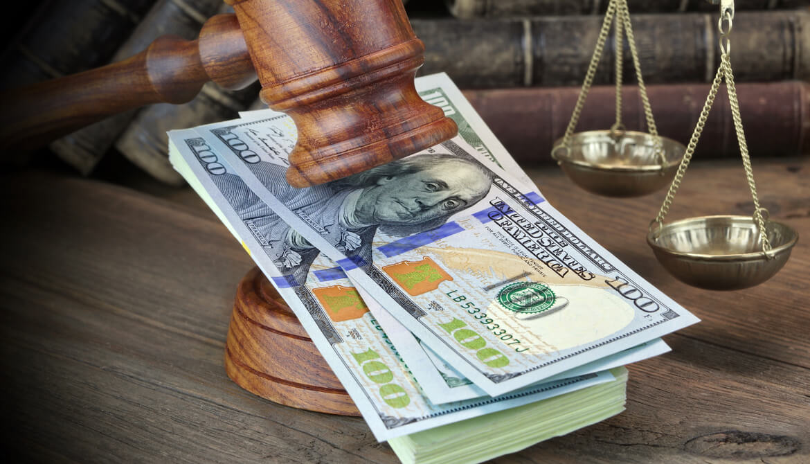 Wooden gavel resting on a stack of $100 bills in front of a set of brass scales representing compensation and regulation