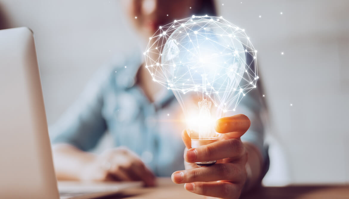 woman at desk holding out a glowing lightbulb with a network of data points forming the image of a brain on top of it