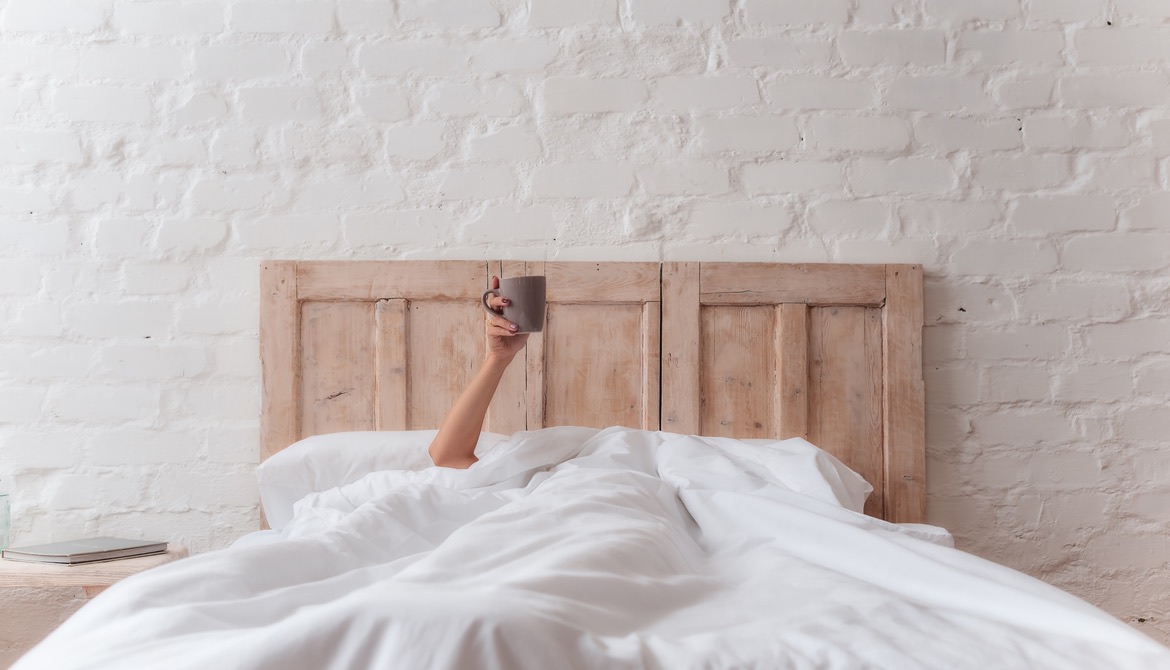 woman’s hand sticking out of white bed covers holding cup of coffee
