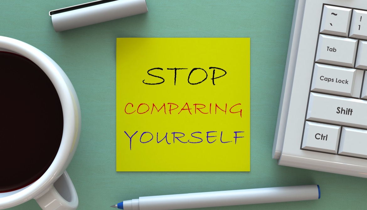 yellow sticky note that says stop comparing yourself on a green desk next to coffee cup and keyboard