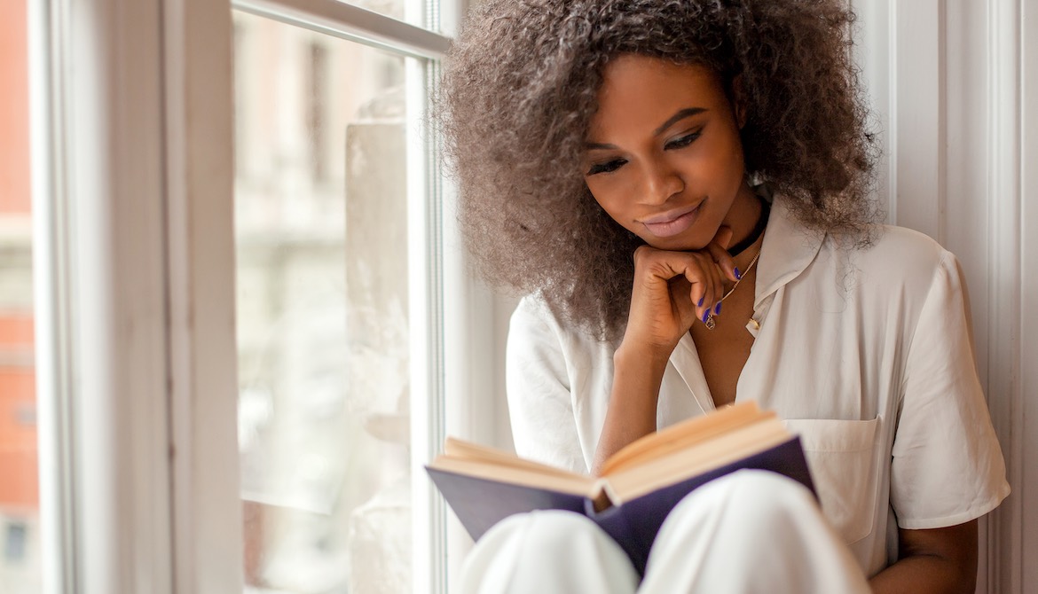 young African American businesswoman reading a book while sitting on window sill
