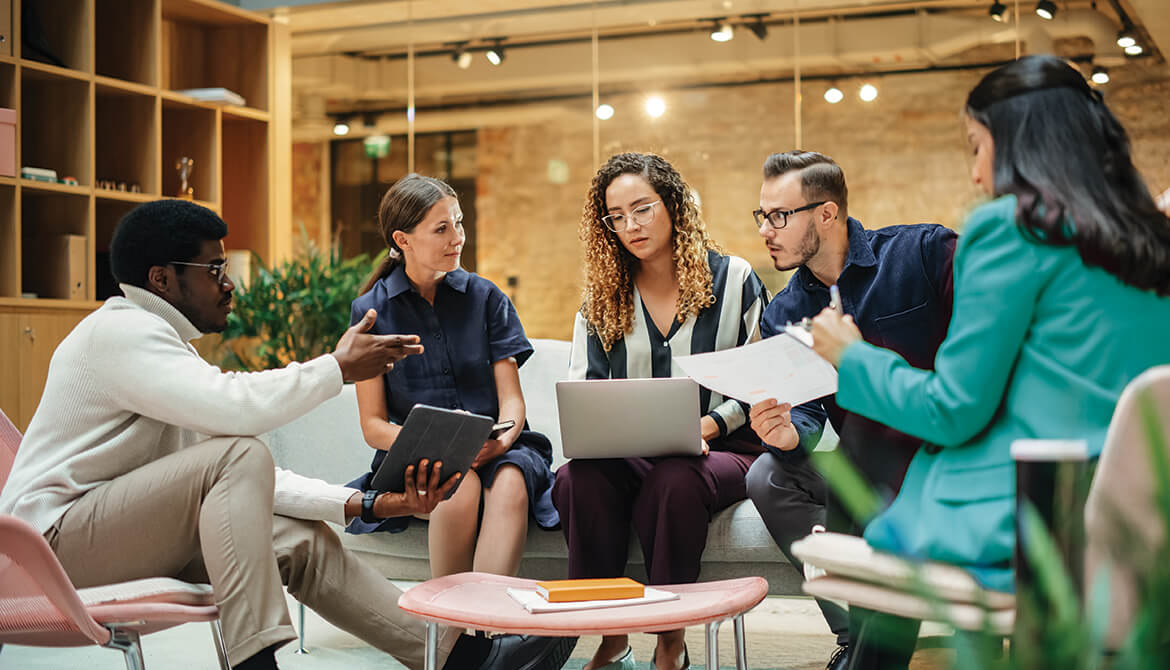 Group of individuals sitting around a coffee table looking at laptops and papers having a meeting