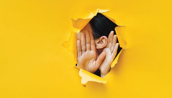 woman holding cupping hands around ear to listen through hole in yellow paper background