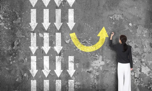 woman in front of black board with lots of white arrows pointing straight down and one yellow arrow curving around to point up, representing making a different decision