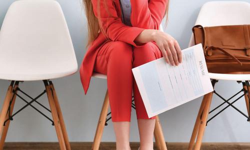 Woman in a red pantsuit sits on a white chair holding her resume while she waits for a job interview