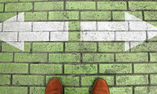 person in brown dress shoes standing on a green brick crossroad with white arrows pointing left and right