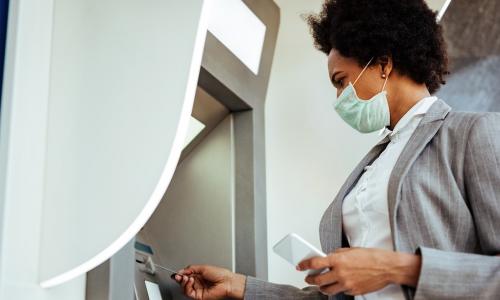 African-American businesswoman wearing mask uses ATM and mobile app