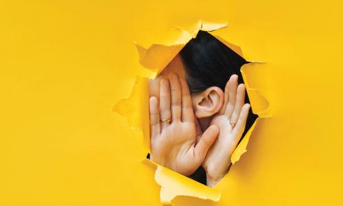 woman holding cupping hands around ear to listen through hole in yellow paper background