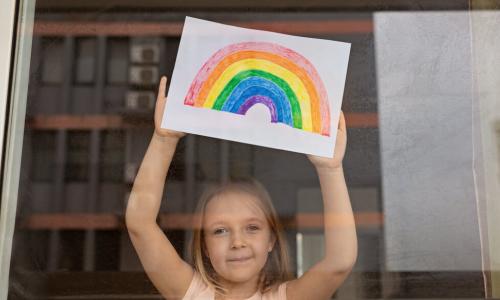 child in window holding up colored drawing of rainbow of hope