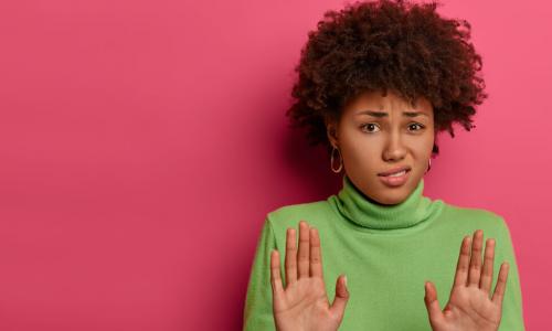 black woman in a green sweater on a pink wall putting her hands up, rejecting something