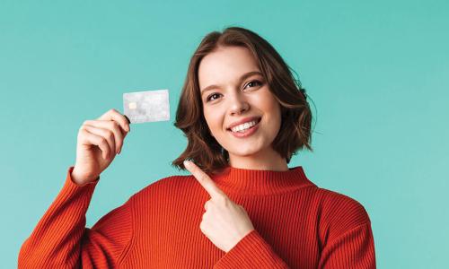 Smiling young woman in orange sweater holding up and pointing to a credit card