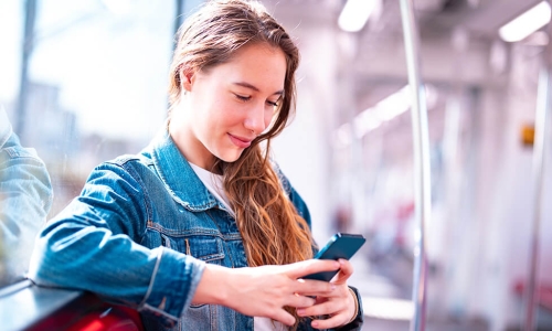 young woman looking at her smart phone while leaning on wall with window behind her