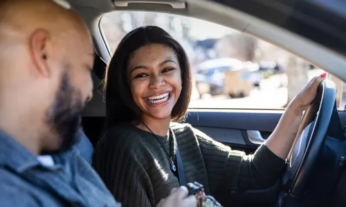 smiling woman sitting in driver seat handing the keys to male passenger in a car